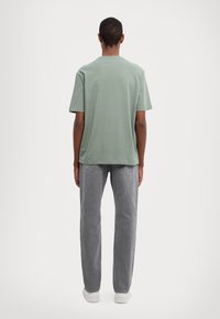 Zelené bavlněné tričko s volným střihem a krátkým rukávem; v kombinaci se šedými rovnými džínami a bílou obuví, pohled zezadu.