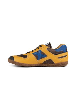 Zapatilla amarilla y marrón con detalle de cruz azul, cordones negros, suela marrón texturizada y parte superior de malla, mostrada desde el lado exterior.