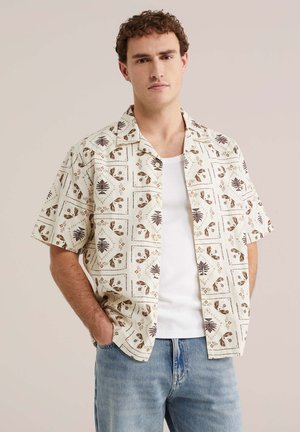 Jeune homme aux cheveux bouclés portant une chemise à manches courtes à motifs boutonnée sur un débardeur blanc et un jean bleu clair, les mains dans les poches.