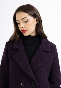 Dobbeltbrystet, lilla uldfrakke med en struktureret overflade, store reverser og to sorte knapper, båret over en sort turtleneck.