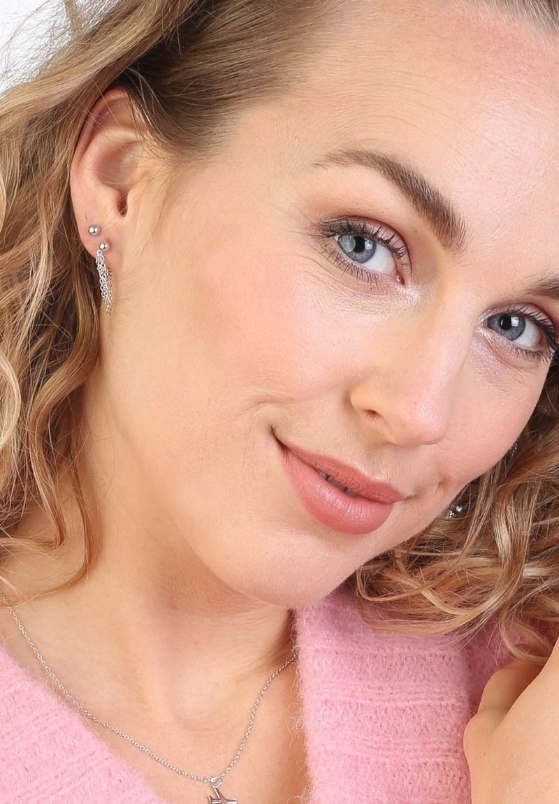 Zilveren oorbellen hebben een delicate, kanten uitstraling met een kleine stud en een druppelvormig element. Het model draagt een zachtroze trui en een zilveren ketting.
