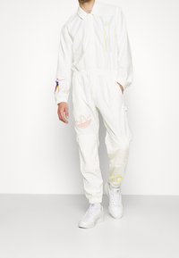 Man in een witte Adidas trainingspak met kleurrijke menselijke figuurontwerpen en witte Adidas sneakers, staand tegen een witte achtergrond.