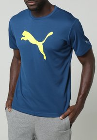 Herrarnas blåa atletiska t-shirt är gjord av ett lättviktigt material. Har en stor gul Puma-logga på framsidan och korta ärmar.