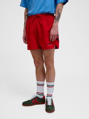 Man draagt rode Hummel sportshorts, witte sokken met groene en rode strepen en groene sneakers met rode accenten, staand tegen een effen achtergrond.