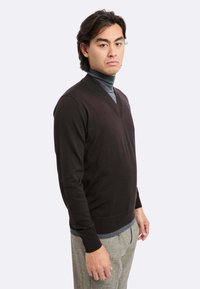 Maglione a V marrone scuro con maniche lunghe e polsini a coste, indossato sopra un dolcevita grigio chiaro, abbinato a pantaloni a quadri.