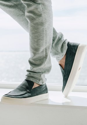 Slip-on sko i mørkegrå ruskind med en hvid gummisål, der har en glat tekstur og et minimalistisk design. Båret med lysegrå stofjoggers.