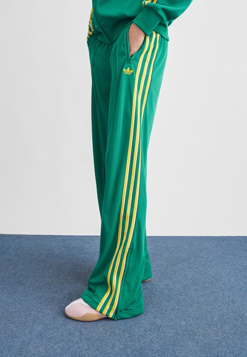 Άτομο που στέκεται φορώντας πράσινο φόρμας με κίτρινες ρίγες και λογότυπο, με τα χέρια στις τσέπες, σε μπλε χαλιά δάπεδο.