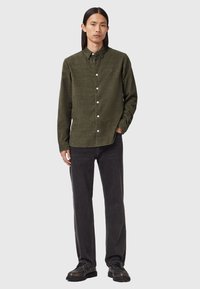 Mand med langt mørkt hår iført en olivengrønn skjorte med knapper, sorte jeans og sorte snørestøvler, står foran en ensfarvet lys baggrund.