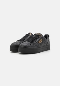 Sorte quiltede sneakers med platformssål, der har sorte snørebånd, guld accenter og "GUESS" branding på pløsen. Glat tekstur.
