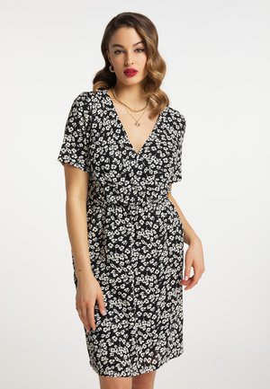 Vrouw draagt een zwarte jurk met witte bloemenprint, V-hals, korte mouwen en gouden kettinghalskettingen tegen een effen witte achtergrond.