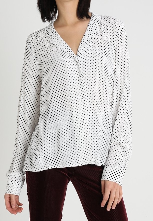 Långärmad blus i vitt tyg med v-ringning och svarta prickar. Designen har en avslappnad passform och subtil textur.