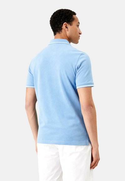 Homme portant un polo bleu clair à manches courtes avec bordure blanche et un pantalon blanc, debout, de dos sur un fond uni.