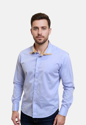 Uomo con capelli castani corti e barba, che indossa una camicia azzurro chiaro con dettagli gialli sul colletto interno, in piedi su uno sfondo bianco.