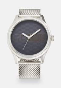 Silberne Metalluhr mit einem Mesh-Armband, dunkelgraues Ziffernblatt mit geprägt eingraviertem "GUESS"-Logo, silberne Zeiger und geriffelter Lünette.
