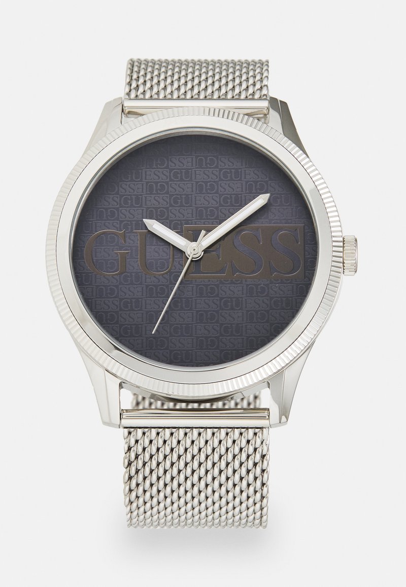 Silberne Metalluhr mit einem Mesh-Armband, dunkelgraues Ziffernblatt mit geprägt eingraviertem "GUESS"-Logo, silberne Zeiger und geriffelter Lünette.