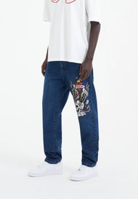 Jeans de denim azul con un corte relajado, que presentan un colorido diseño bordado de una calavera y una serpiente en el muslo izquierdo. Llevados con una camiseta blanca.