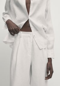Chemise en lin blanc à boutons, légèrement déboutonnée à la taille, associée à un pantalon taille haute assorti, mettant en valeur une texture douce et des lignes épurées.