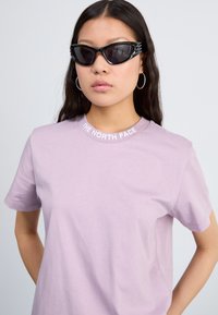 Camiseta de color morado claro con cuello redondo que presenta el logo de "THE NORTH FACE" en blanco. El modelo lleva gafas de sol negras de estilo cat-eye y pendientes de aro.
