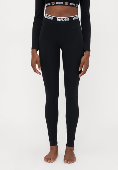 Fekete Moschino leggings-t és hozzá illő, logós derékpántos cropped topot viselő nő, mezítláb áll a fehér háttér előtt.