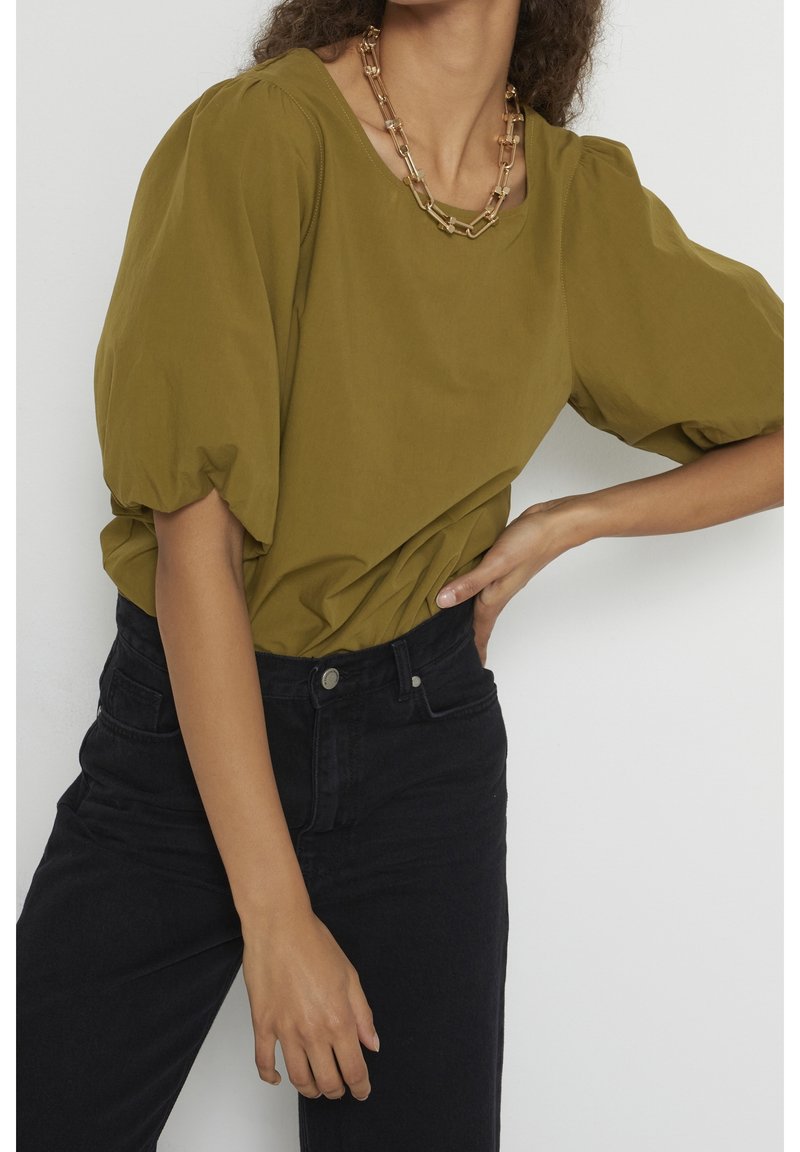 Blouse olive à manches bouffantes en tissu doux, associée à un jean noir taille haute. Un collier en chaîne dorée ajoute une touche métallique.