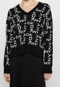 Musta neulepaita, jossa on V-pääntie, ja joka esittelee toistuvaa valkoista "DKNY"-logoa. Laajentuva helma ja pitkät hihat, joissa on ribbireunat.