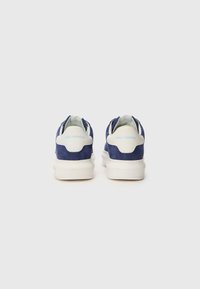 Sneakers de camurça azul marinho com sola de borracha branca e marcação azul clara no calcanhar. Design arredondado, estilo minimalista e silhueta de cano baixo.