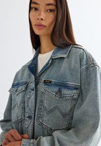 Νέα γυναίκα με μακριά σκούρα μαλλιά, φορώντας φθαρμένο μπλε τζιν μπουφάν πάνω από λευκό τοπ, με τα χέρια της σταυρωμένα μπροστά, ουδέτερο φόντο.