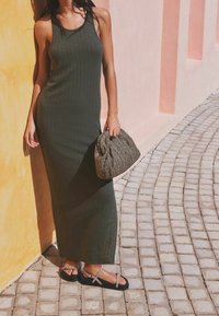 Vrouw in een mouwloze maxi-jurk in olijfgroen staat op een geplaveid pad en houdt een geweven tas vast bij gele en roze muren.