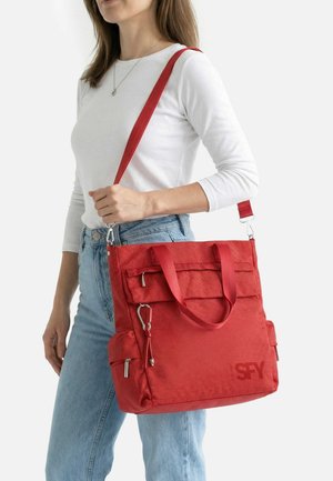 Donna con top bianco e jeans blu che porta una grande borsa a tracolla rossa con molteplici tasche con cerniera e tracolla staccabile.