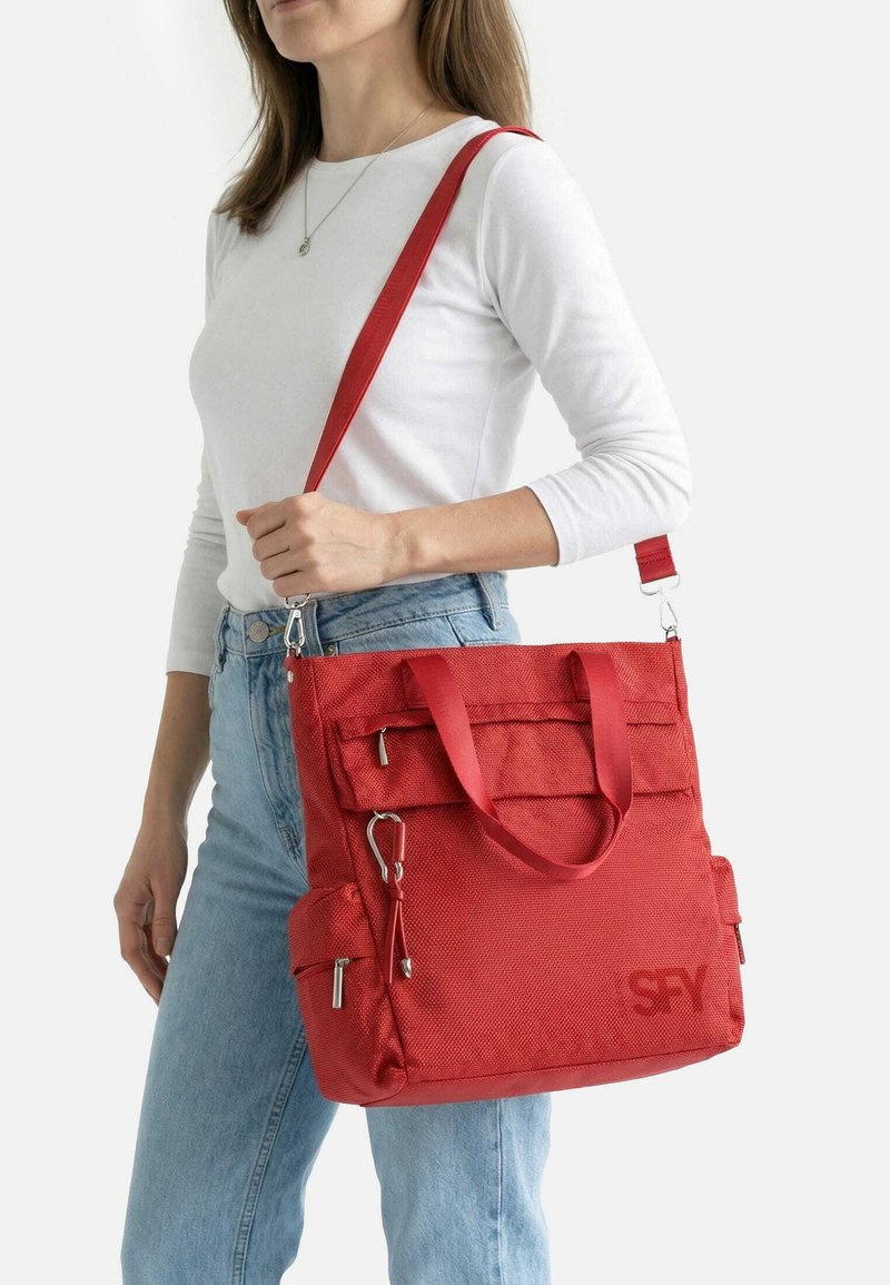 Woman wearing white top and blue jeans carries large red shoulder bag with multiple zippered pockets and a detachable strap.