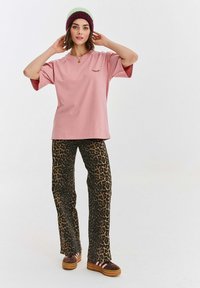 Světle růžové tričko s krátkým rukávem a grafikou řas, v kombinaci s hnědými kalhotami s leopardím potiskem a hnědými platformovými botami s růžovými pruhy.