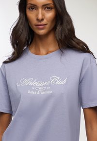 T-shirt cotone lavanda con collo rotondo, con testo ricamato bianco: "Athleisure Club 1993 NY Relax & Wellness." Maniche corte.