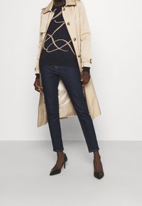 Beige trenchcoat med bruna detaljer, marinblå tröja med guldmönster, mörkblå skinny jeans och svarta spetsiga klackar.