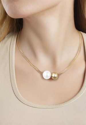 Halskæde med to store perler, en hvid perle-lignende og en guldfarvet, båret på en tynd guldkæde rundt om en persons hals.