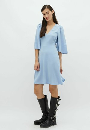 Robe bleu clair avec décolleté en V et manches évasées, associée à des bottes noires hauteur genou avec des accents de boucles. Texture lisse, silhouette fluide.