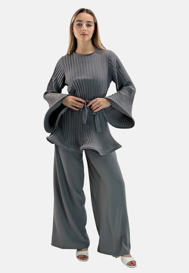 Top gris plisado con mangas acampanadas y un lazo en la cintura, combinado con pantalones anchos gris. La tela tiene una textura sedosa, ofreciendo una silueta fluida.