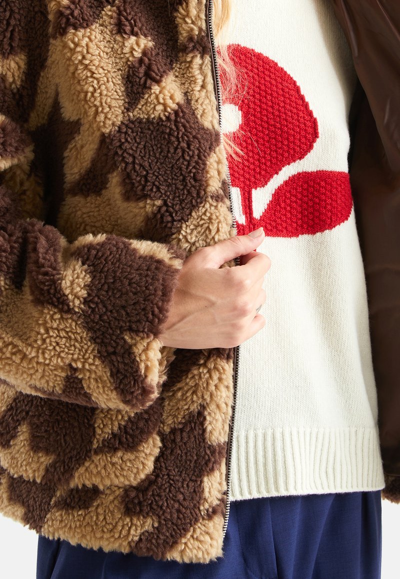 Braune und beige gemusterte Fleecejacke mit Reißverschluss, kombiniert mit einem cremefarbenen Pullover mit rotem Grafikdesign. Weicher Stoff.