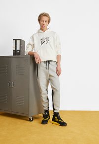 Lichtgrijze sweatpants met zwarte zijstreep, gecombineerd met een crème hoodie met een zwart bliksemschichtgrafiek. Zwart-en-gele sportieve sneakers.