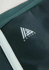 Træningstøj i mørkegrønt stof med en struktureret overflade. Har et hvidt logo og en kontrasterende lysegrøn kant langs kanten.