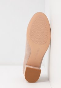 Chaussure en daim beige unique avec une semelle marron texturée et un petit talon carré, positionnée sur un fond d'angle blanc.