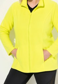 Kirkas keltainen fleece-takki, jossa on vetoketjukiinnitys, sivutaskut ja pystykaulus. Yhdistetty mustiin legginsseihin. Sujuva pinta.