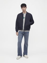 Jeune homme debout portant une veste noire, un sweat gris, un jean bleu et des baskets blanches sur un fond blanc uni.