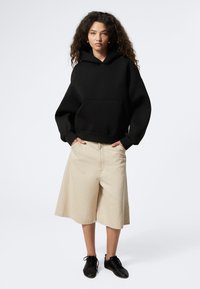 Svart huvtröja med ficka framtill, i kombination med beige shorts som slutar vid knäna. Modellen bär svarta skor. Flat och slät tygtextur.