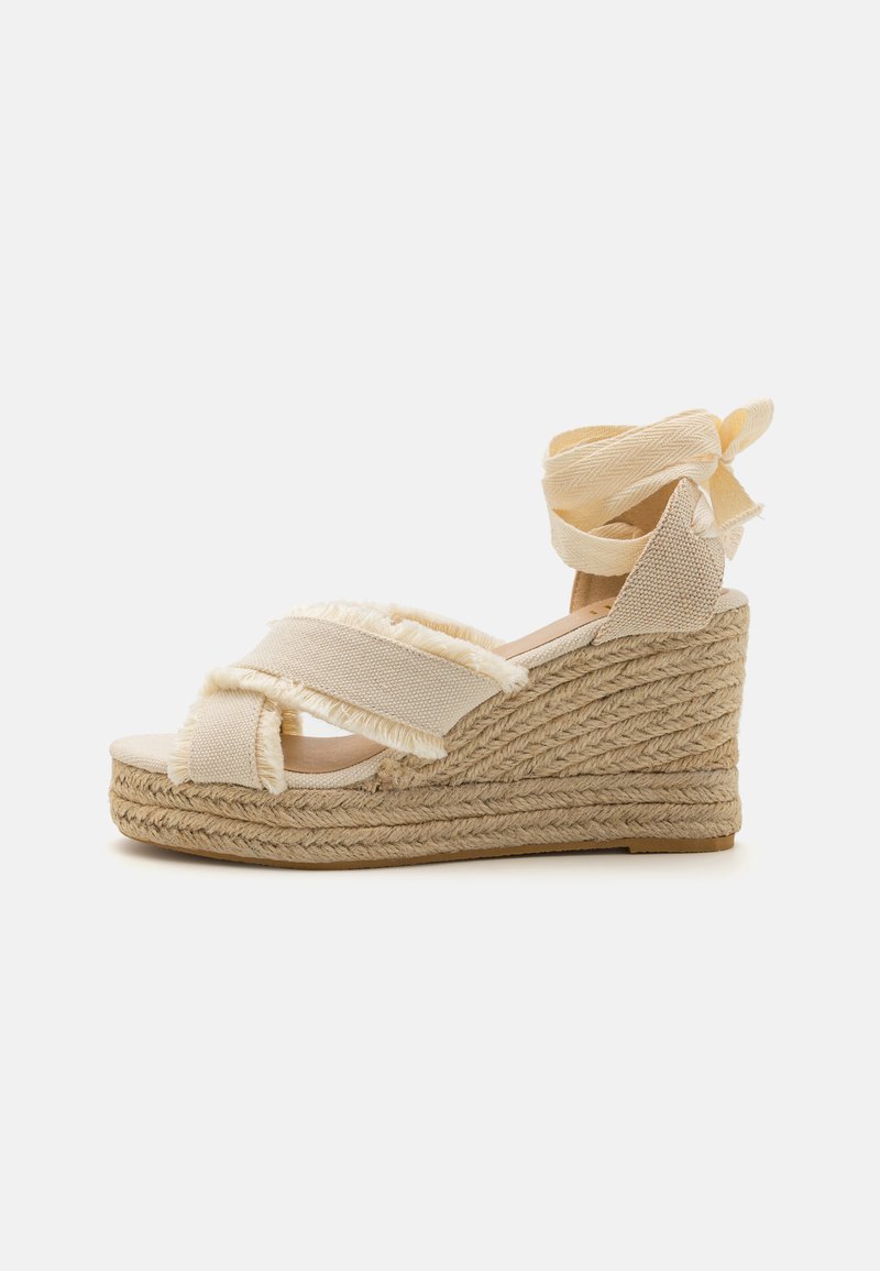 Sandalia de cuña de esparto beige con correas de tela deshilachadas, suela de yute trenzado y un acabado texturizado. Con un lazo de cinta en el tobillo.