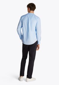 Lys blå langermet skjorte med knappede mansjetter og avrundet kant, brukt med svarte bukser og hvite joggesko. Synlig glatt stofftekstur.