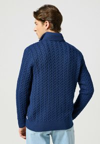 Granatowy sweter z dzianiny z wysokim kołnierzem, fakturowanym wzorem w warkocze, ściągaczami przy mankietach i u dołu. Akcentowany małą metką z logo po boku.