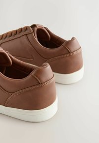 Een paar bruine leren sneakers met witte zolen, gezien vanuit de achterkant, tentoongesteld op een lichte ondergrond.