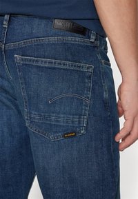 Acercamiento al área del bolsillo trasero de unos jeans de mezclilla azul con la etiqueta del logo de G-Star y un parche de cuero negro de la marca encima del bolsillo, combinados con una camisa azul marino.