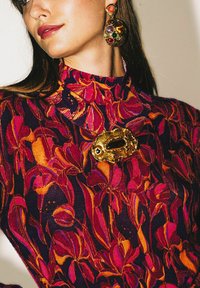 Blusa con motivo floreale caratterizzata da disegni floreali viola intenso e arancione vivace, arricchita da una spilla dorata lavorata e orecchini ornati.