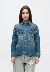 Νέα γυναίκα που φοράει μπλε τζιν μπουφάν με διακοσμητικά από λουλουδένιες στρας και ασημένιο μεταλλικό παντελόνι, στέκεται μπροστά σε ένα απλό λευκό φόντο.